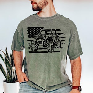 May include: A green t-shirt with a black and white graphic of an off-road vehicle driving over an American flag.