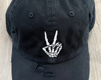 May include: Black distressed baseball cap with a white embroidered skeleton hand making the peace sign.