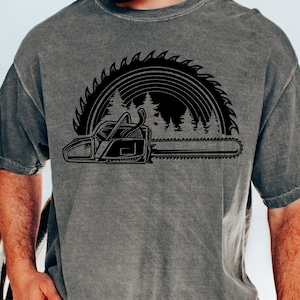 May include: A gray t-shirt with a black graphic of a chainsaw and a saw blade with a forest scene in the background.