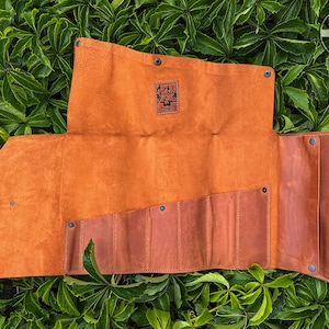 Puede incluir: Un rollo de herramientas de cuero marrón con seis ranuras para herramientas. El rollo está desenrollado y tiene una correa de cuero con una hebilla de metal. El rollo está etiquetado como "Fadir Tools Est. 2016".