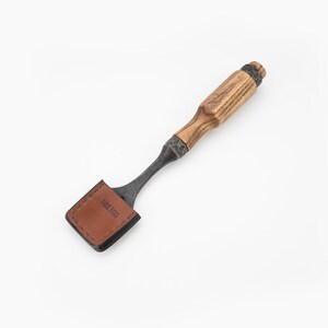 May include: A brown leather-wrapped chisel with an oak wood handle and a metal blade. The handle has a dark brown finish. The blade is made of steel and has a black finish. The chisel is labelled "Habit Tools".