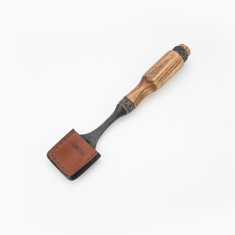 May include: A brown leather-wrapped chisel with an oak wood handle and a metal blade. The handle has a dark brown finish. The blade is made of steel and has a black finish. The chisel is labelled "Habit Tools".