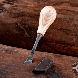 May include: A small, wooden-handled carving tool with a metal blade. The tool is lying on a wooden surface. The handle is light brown with dark brown streaks. The blade is dark gray and has a slightly curved shape. The tool is made by Fadir Tools, EST. 2016.