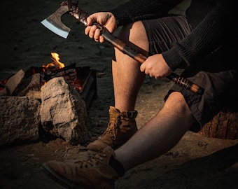 May include: A person holding a brown-handled axe with a leather wrap and a silver blade. The axe is held in front of a campfire with rocks and burning wood.