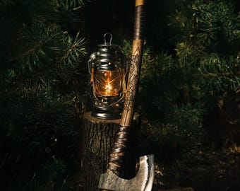 May include: A hand-forged axe with a wooden handle and leather wrap. The handle is decorated with intricate carvings. The axe is resting on a tree stump next to a lantern.