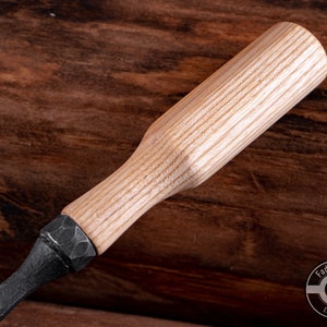 May include: A close-up of a wooden handled carving tool with a black metal blade. The handle is made of light wood with a natural grain pattern. The tool is resting on a wooden surface.