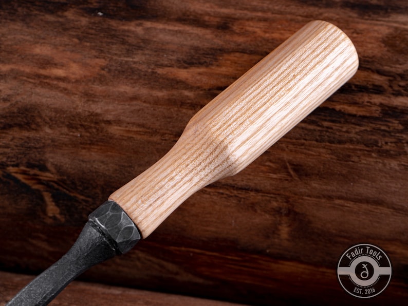 May include: A close-up of a wooden handled carving tool with a black metal blade. The handle is made of light wood with a natural grain pattern. The tool is resting on a wooden surface.