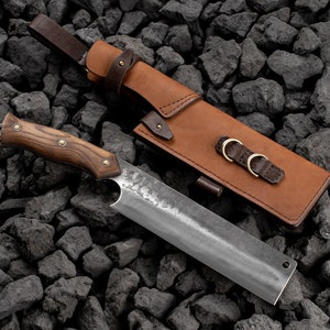 May include: A large, handmade knife with a wooden handle and a leather sheath. The knife has a textured blade and is resting on a bed of black rocks. The sheath has two metal buckles and a brown leather strap.