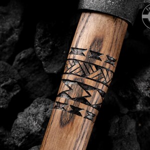 Puede incluir: Primer plano del mango de un hacha de madera con intrincados patrones geom&eacute;tricos tallados. El mango es de color marr&oacute;n claro con vetas oscuras. La cabeza del hacha es de metal oscuro. El logotipo "Fadir Tools EST. 2016" es visible.