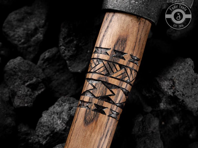 Puede incluir: Primer plano del mango de un hacha de madera con intrincados patrones geom&eacute;tricos tallados. El mango es de color marr&oacute;n claro con vetas oscuras. La cabeza del hacha es de metal oscuro. El logotipo "Fadir Tools EST. 2016" es visible.