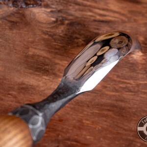 May include: A close-up of a wood carving tool with a curved blade and a wooden handle. The blade is polished and reflects the surrounding wood.