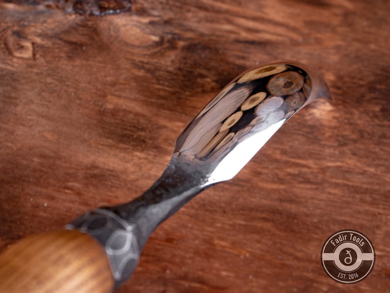 May include: A close-up of a wood carving tool with a curved blade and a wooden handle. The blade is polished and reflects the surrounding wood.