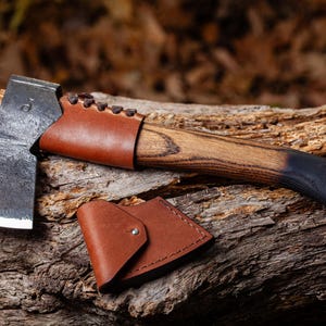 May include: A handcrafted axe with a dark, weathered blade and a wooden handle. The handle transitions from a natural wood tone to black. A brown leather sheath and handle cover are also included. The axe rests on a piece of wood.