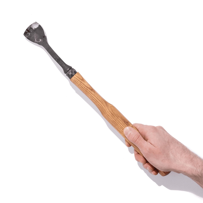 May include: A hand holds a wooden-handled tool with a metal head. The tool has a curved, spoon-like shape at the end.