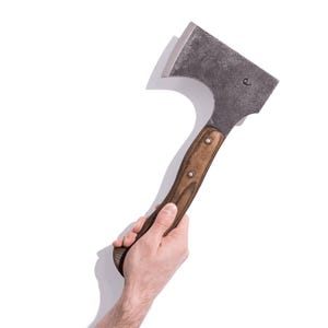 May include: A metal axe with a wooden handle. The axe head is dark gray, and the handle is brown with two rivets. The axe is held in a person's hand against a white background.
