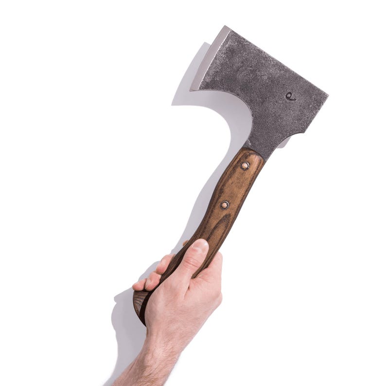 May include: A metal axe with a wooden handle. The axe head is dark gray, and the handle is brown with two rivets. The axe is held in a person's hand against a white background.