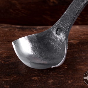 May include: A close-up of a silver metal spoon with a rounded bowl. The spoon is resting on a wooden surface. The spoon has a small mark on the back of the bowl.
