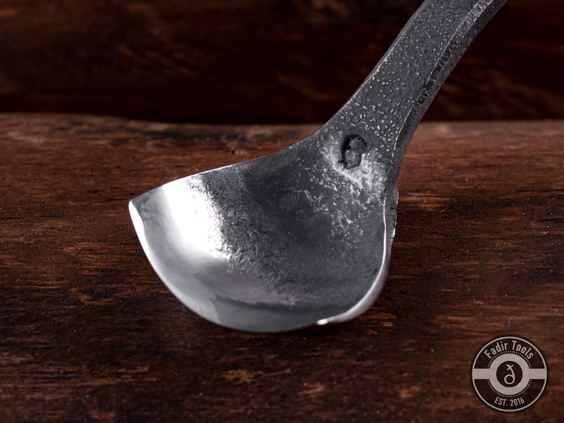May include: A close-up of a silver metal spoon with a rounded bowl. The spoon is resting on a wooden surface. The spoon has a small mark on the back of the bowl.