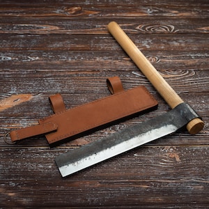 May include: A brown leather sheath with a metal hook and a wooden handle attached to a long, curved, metal blade. The blade is dull and has a slight curve. The sheath and blade are lying on a wooden surface.