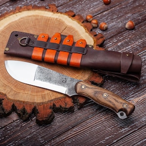 Puede incluir: Un cuchillo de caza artesanal con mango de madera y funda de cuero. El cuchillo tiene una hoja de acero inoxidable y está sobre una superficie de madera. La funda es marrón con correas de cuero naranja.