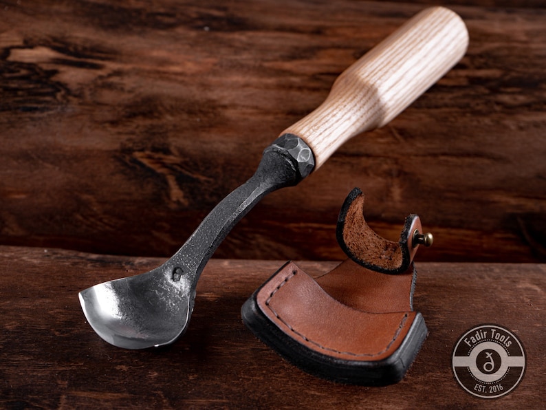 May include: A wooden handled spoon carving tool with a curved metal blade. The tool is laying on a wooden surface next to a brown leather sheath.