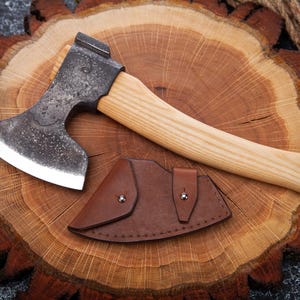 May include: A small, hand-forged axe with a wooden handle and a leather sheath. The axe is resting on a wooden surface.