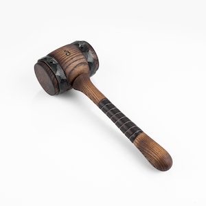 May include: A wooden gavel with a dark brown head and a lighter brown handle. The head is encircled by a black metal band. The handle features black leather bands for grip. The gavel is set against a white background.