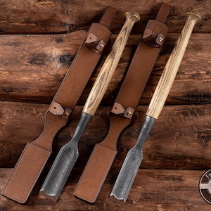 Könnte beinhalten: Drei Holz-Meißel mit Lederscheiden. Die Meißel haben Metallklingen und Holzgriffe mit einem gestreiften Muster. Die Scheiden sind aus braunem Leder mit einem genähten Design. Die Meißel sind in einer Reihe auf einer Holzoberfläche angeordnet. Der Text "Fadir Tools Est. 2016" ist in der unteren rechten Ecke des Bildes sichtbar.