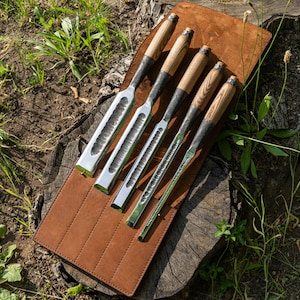 Puede incluir: Conjunto de cinco cinceles para tallar madera con mangos de madera, dispuestos en un rollo de cuero marrón. Los cinceles tienen diferentes tamaños y formas.