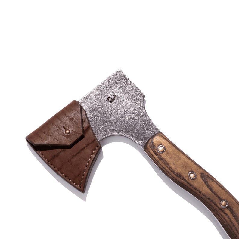 May include: A small axe with a wooden handle and a metal head. The axe head has a leather sheath. The handle is made of wood and is attached to the metal head with rivets. The axe is designed for chopping wood.