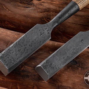 May include: Two black metal chisels with wooden handles, lying on a wooden surface. The chisels have a textured finish and are labeled with the letter "P".
