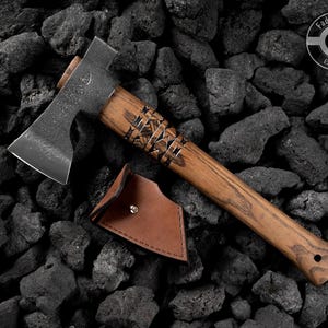 Puede incluir: Un hacha artesanal con mango de madera y hoja de metal, sobre rocas gris oscuro. El mango tiene tallas decorativas. Una funda de cuero marr&oacute;n est&aacute; junto al hacha. El texto "Fadir Tools EST. 2016" est&aacute; en el hacha.