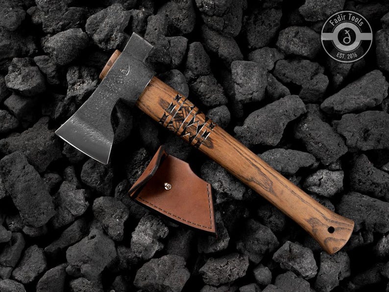 Puede incluir: Un hacha artesanal con mango de madera y hoja de metal, sobre rocas gris oscuro. El mango tiene tallas decorativas. Una funda de cuero marr&oacute;n est&aacute; junto al hacha. El texto "Fadir Tools EST. 2016" est&aacute; en el hacha.
