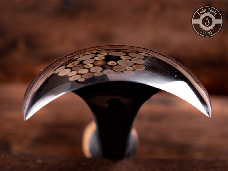 May include: A close-up of a curved, polished metal woodworking tool with a wooden handle. The tool is designed for carving and shaping wood. The metal surface reflects the surrounding environment, showing a pattern of small, round shapes.