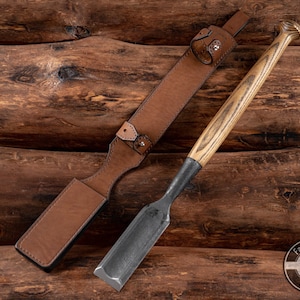 Puede incluir: Una funda de cuero marrón con cierre de presión metálico y un cincel con mango de madera y hoja de metal negro. Las herramientas están sobre un fondo de madera con el texto "Fadir Tools Est. 2016" en un círculo.