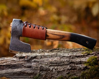 May include: A small, hand-forged axe with a brown leather handle wrap and a wooden handle with a black finish. The axe is resting on a piece of weathered wood.