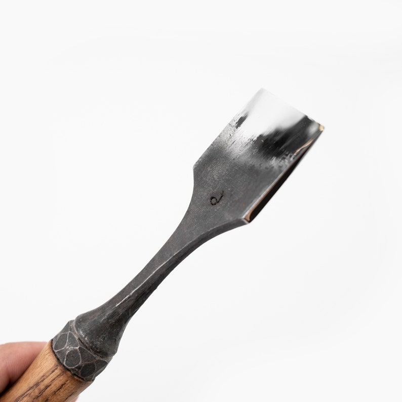 May include: A close-up of a wood-handled chisel with a metal blade. The blade is rectangular and has a slight curve. The handle is made of wood and has a dark brown finish. The chisel is marked with the number "6" on the blade.
