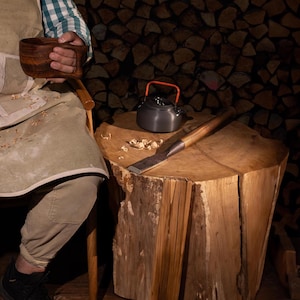 Puede incluir: Una mesa de tocón de árbol con una tetera de metal, un cuenco de madera y un cincel de madera. La mesa está hecha de una sola pieza de madera y tiene un aspecto natural y rústico.
