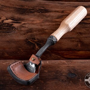 May include: A wood carving tool with a leather sheath and a wooden handle. The tool is made of metal and has a sharp blade. The handle is made of light wood and has a natural finish. The tool is on a wooden surface.