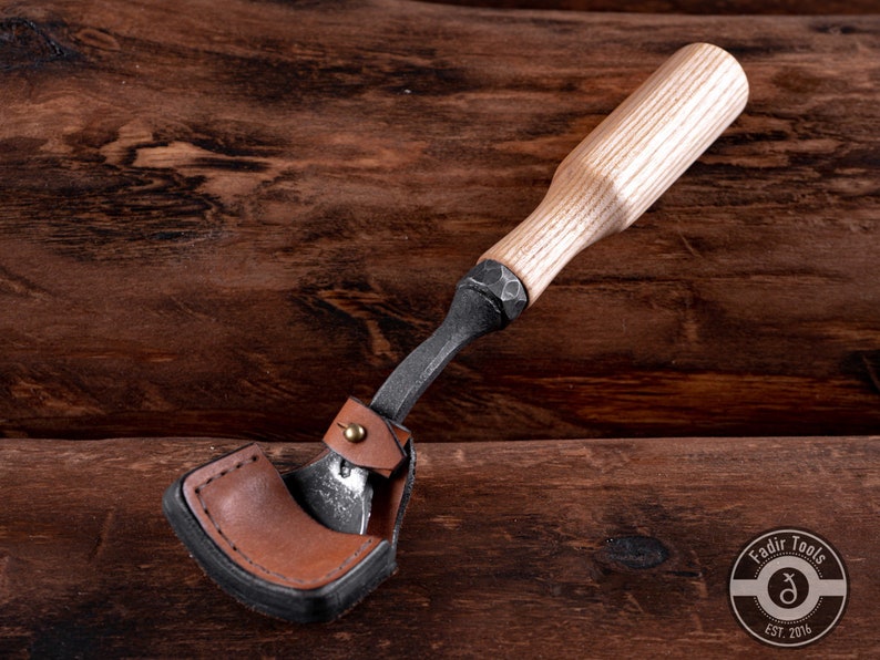 May include: A wood carving tool with a leather sheath and a wooden handle. The tool is made of metal and has a sharp blade. The handle is made of light wood and has a natural finish. The tool is on a wooden surface.