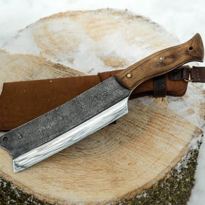 May include: A large, handmade knife with a brown wooden handle and a leather sheath. The blade is made of steel and has a textured finish. The knife is resting on a wooden stump.