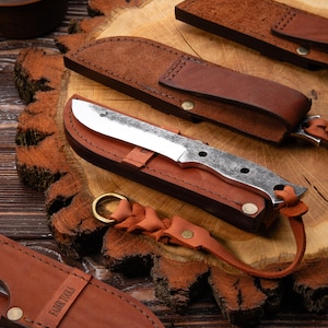 Puede incluir: Colección de cuchillos artesanales con fundas de cuero. Los cuchillos tienen hojas plateadas y mangos texturizados. Las fundas son de cuero marrón con detalles cosidos y correas. Los cuchillos se exhiben sobre una superficie de madera.