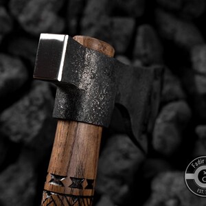 Puede incluir: Primer plano de un hacha artesanal con una hoja oscura y texturizada y una cabeza plateada pulida. El mango de madera presenta dise&ntilde;os tallados. El hacha se encuentra sobre un fondo oscuro y rocoso. El hacha es una herramienta.