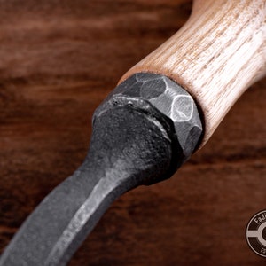 May include: Close-up of a black metal tool with a wooden handle. The handle is made of light-colored wood and has a textured surface. The metal part of the tool is hammered and has a rough finish.