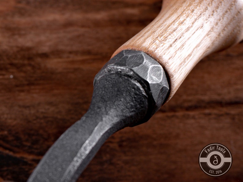 May include: Close-up of a black metal tool with a wooden handle. The handle is made of light-colored wood and has a textured surface. The metal part of the tool is hammered and has a rough finish.