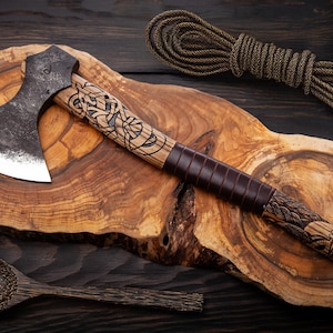 Puede incluir: Un hacha forjada a mano con un mango de madera que presenta un diseño tallado. El hacha está apoyada en una tabla de cortar de madera.
