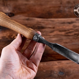 May include: A wood carving tool with a wooden handle and a metal blade. The blade is curved and has a sharp edge. The handle is brown and has a grain pattern. The tool is held in a hand.