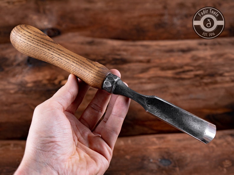 May include: A wood carving tool with a wooden handle and a metal blade. The blade is curved and has a sharp edge. The handle is brown and has a grain pattern. The tool is held in a hand.