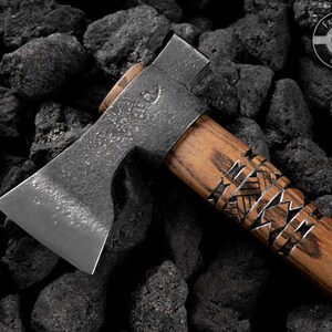 Puede incluir: Un hacha artesanal con un mango de madera con intrincados grabados. La cabeza del hacha es de metal y tiene una hoja afilada. El hacha se encuentra sobre un fondo de rocas oscuras y texturizadas. El logotipo "Fadir Tools EST. 2016" es visible.