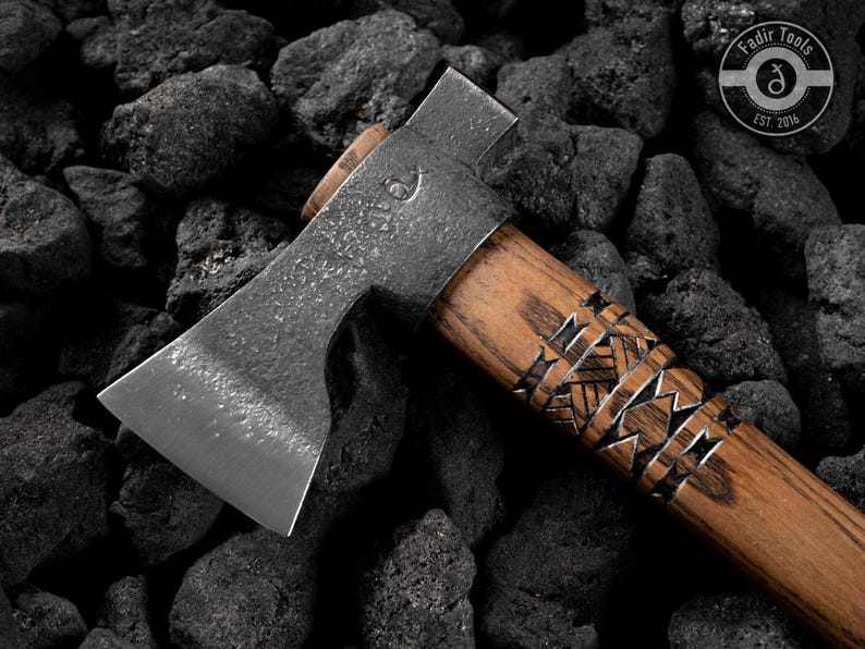 Puede incluir: Un hacha artesanal con un mango de madera con intrincados grabados. La cabeza del hacha es de metal y tiene una hoja afilada. El hacha se encuentra sobre un fondo de rocas oscuras y texturizadas. El logotipo "Fadir Tools EST. 2016" es visible.